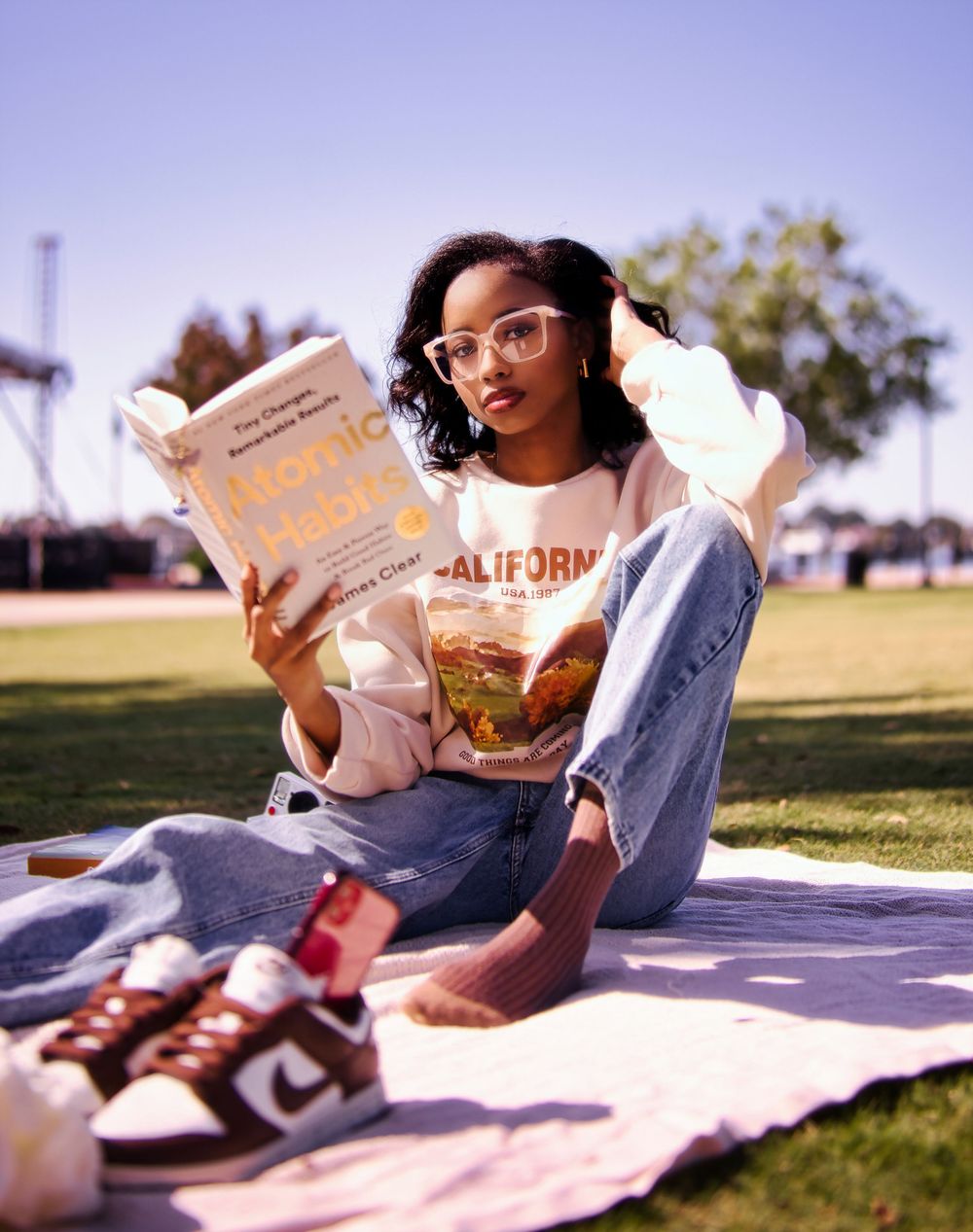 Young woman reading a book on a blanket in a park, wearing a California sweatshirt, jeans, and glasses.