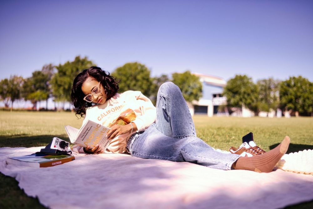 Young woman relaxing on a blanket in a sunny park, reading a book while lying on green grass.