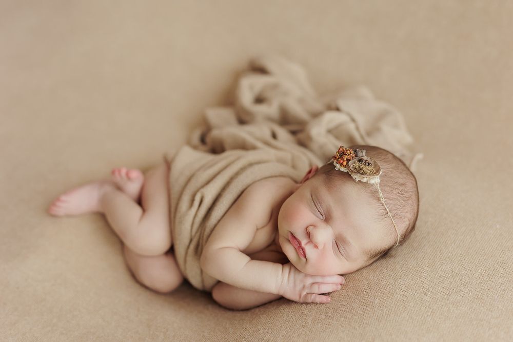 Sleeping newborn wrapped in soft beige fabric with delicate headband resting on neutral backdrop.