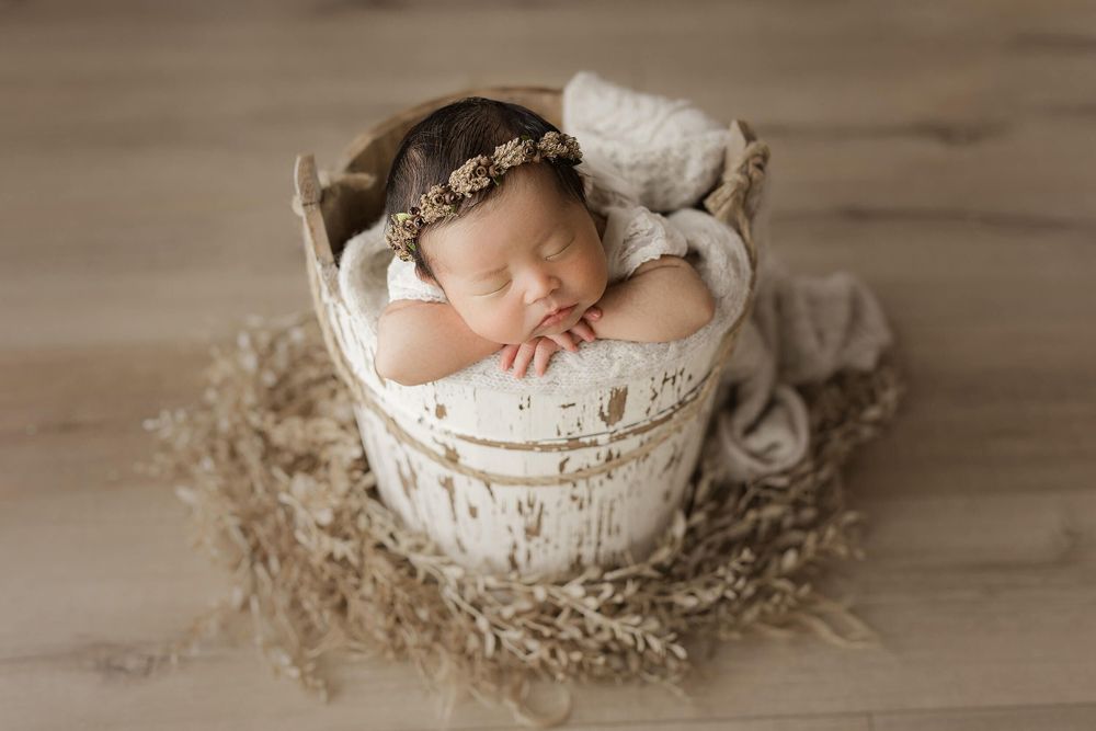Sleeping newborn nestled in white wooden bucket with delicate headband surrounded by dried flowers on neutral background.