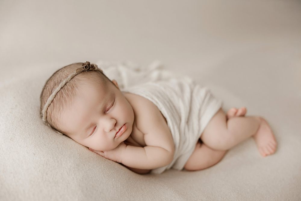 Peaceful sleeping newborn baby wearing white wrap and delicate headband rests on cream colored blanket.
