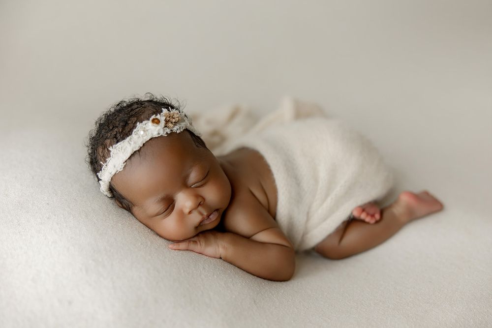 Peaceful sleeping newborn baby wrapped in soft white fabric with delicate lace headband on cream background.