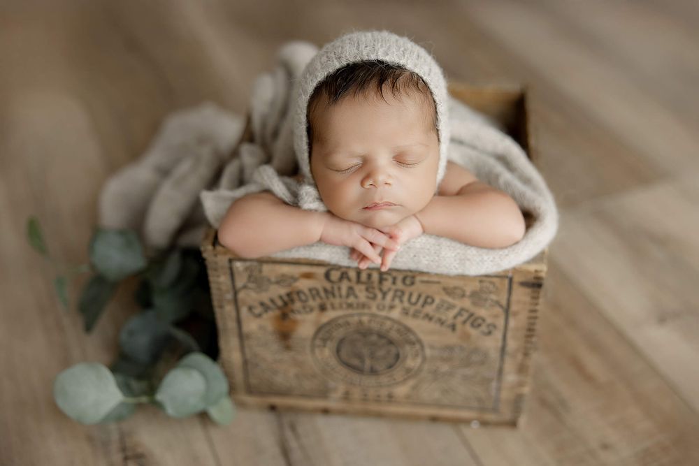 Sleeping newborn rests peacefully in vintage wooden crate wearing white knit bonnet with eucalyptus leaves as decor.