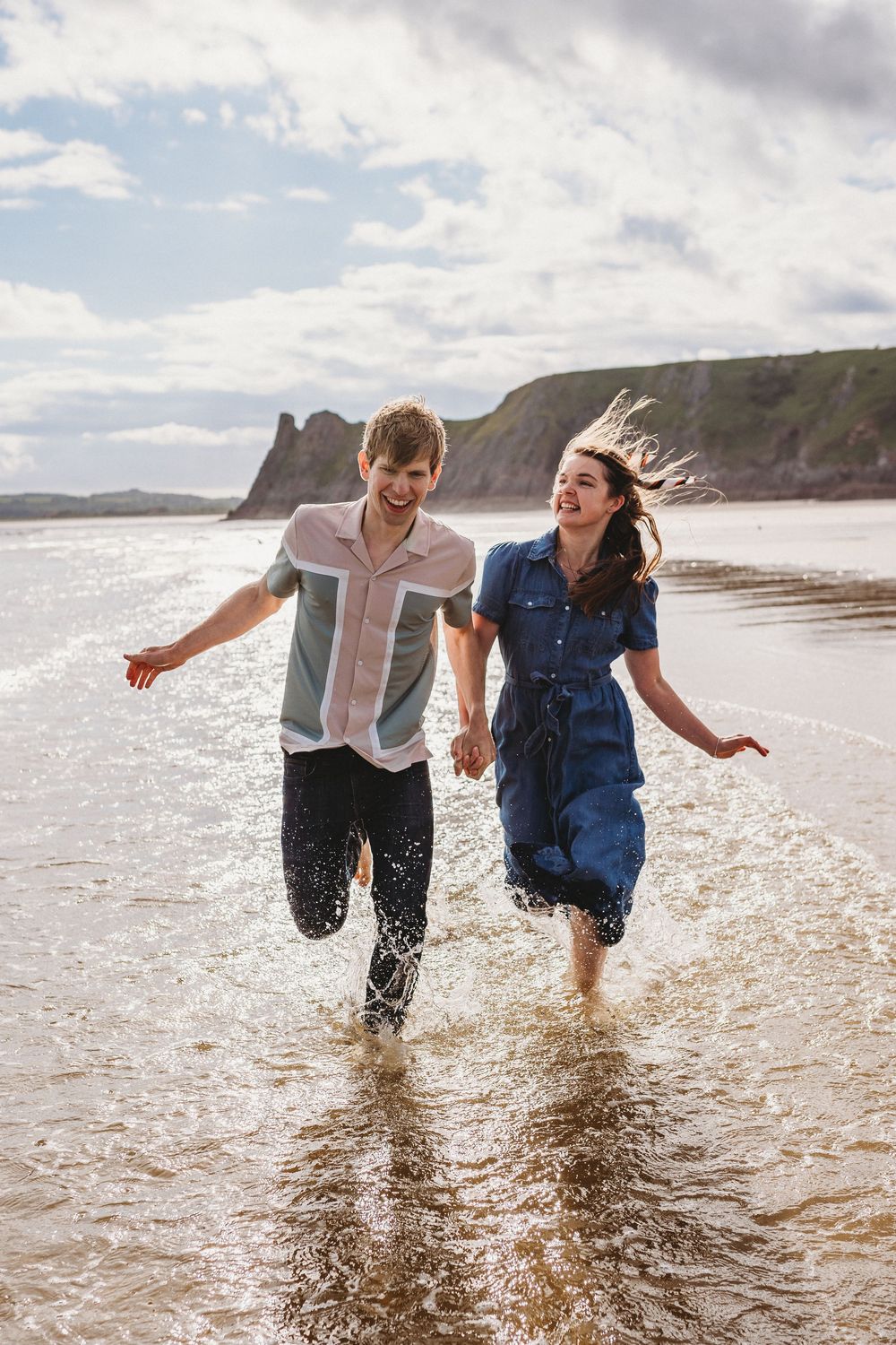 A couple splashes through shallow ocean water along a scenic beach with dramatic cliffs in the background.