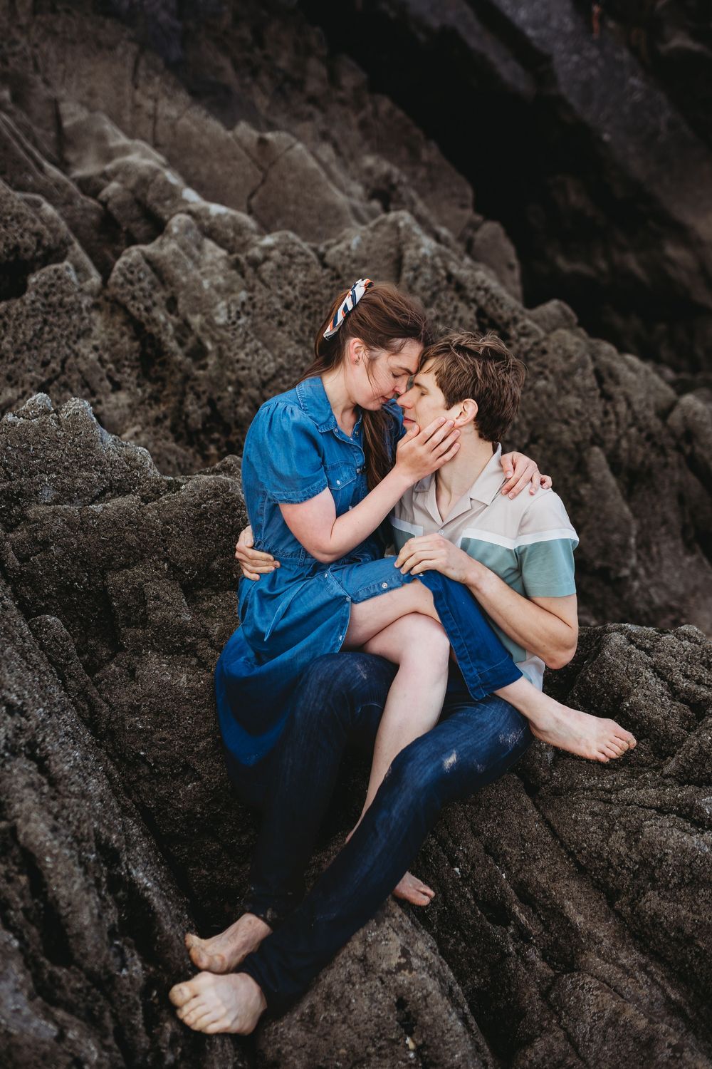 A couple shares intimate moments while sitting on dark rocky coastal terrain at sunset.