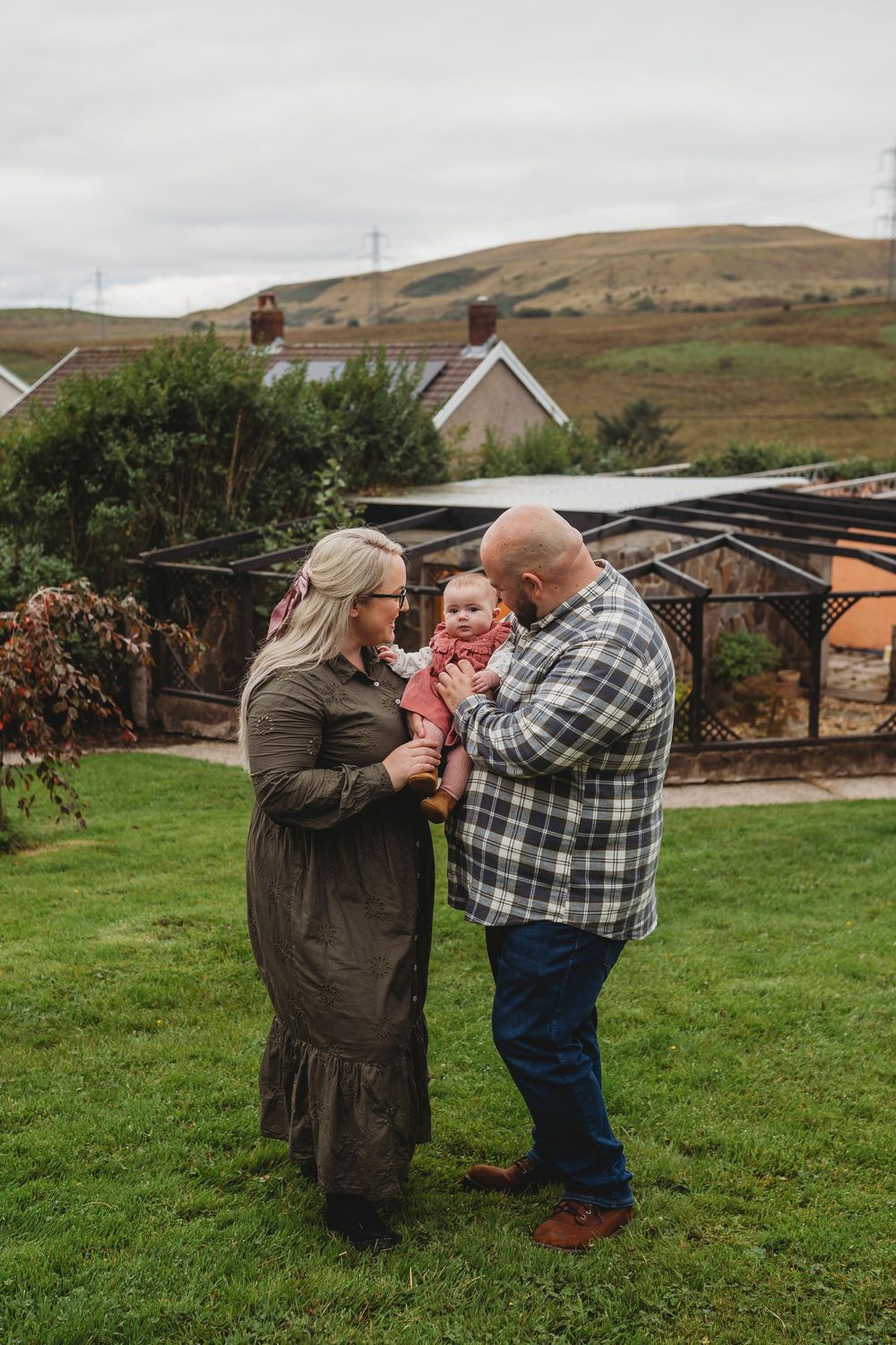 A family shares a tender moment together on a grassy lawn with rolling hills and a greenhouse in the background.