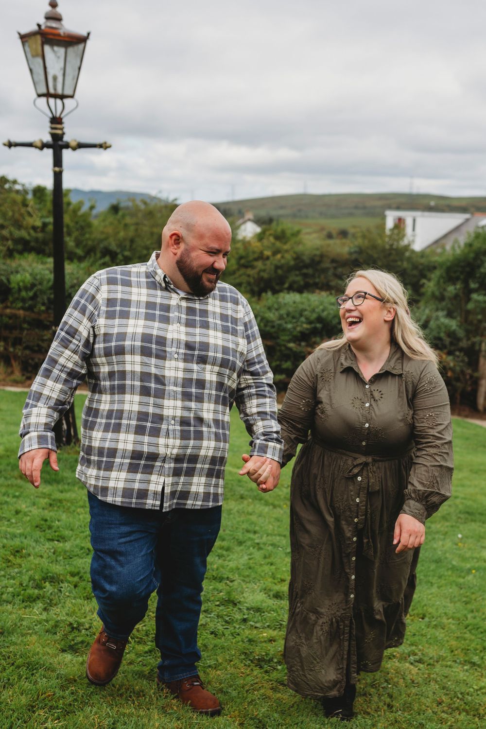 A couple shares a joyful laugh while holding hands and walking on green grass with a lamp post and hills in the background.