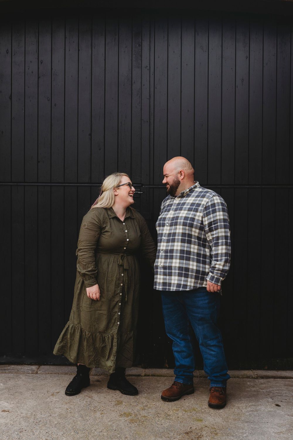 A couple shares a joyful moment together against a black wooden wall, wearing casual fall attire.