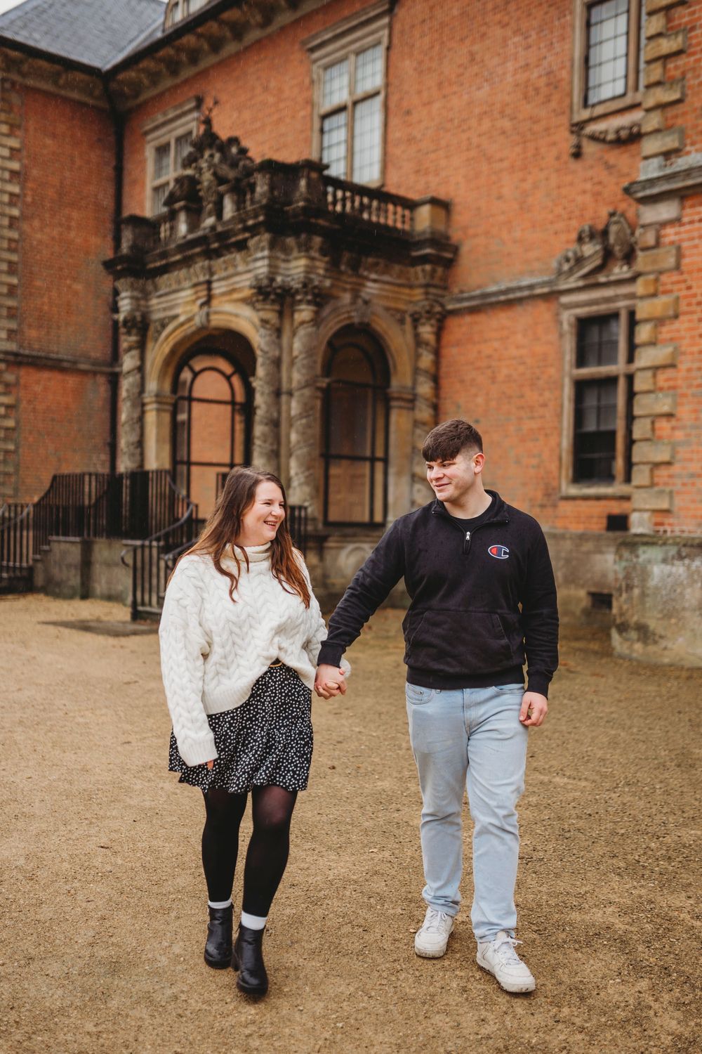 A couple walks together outside a grand brick building with ornate architecture.