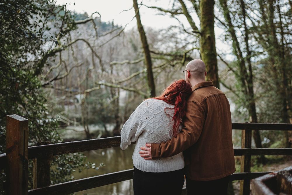 A couple embraces outdoors in a romantic moment while standing on a wooden deck surrounded by trees.