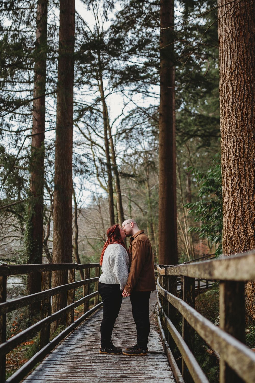 A couple stands together on a wooden boardwalk path surrounded by tall evergreen trees in a forest setting.