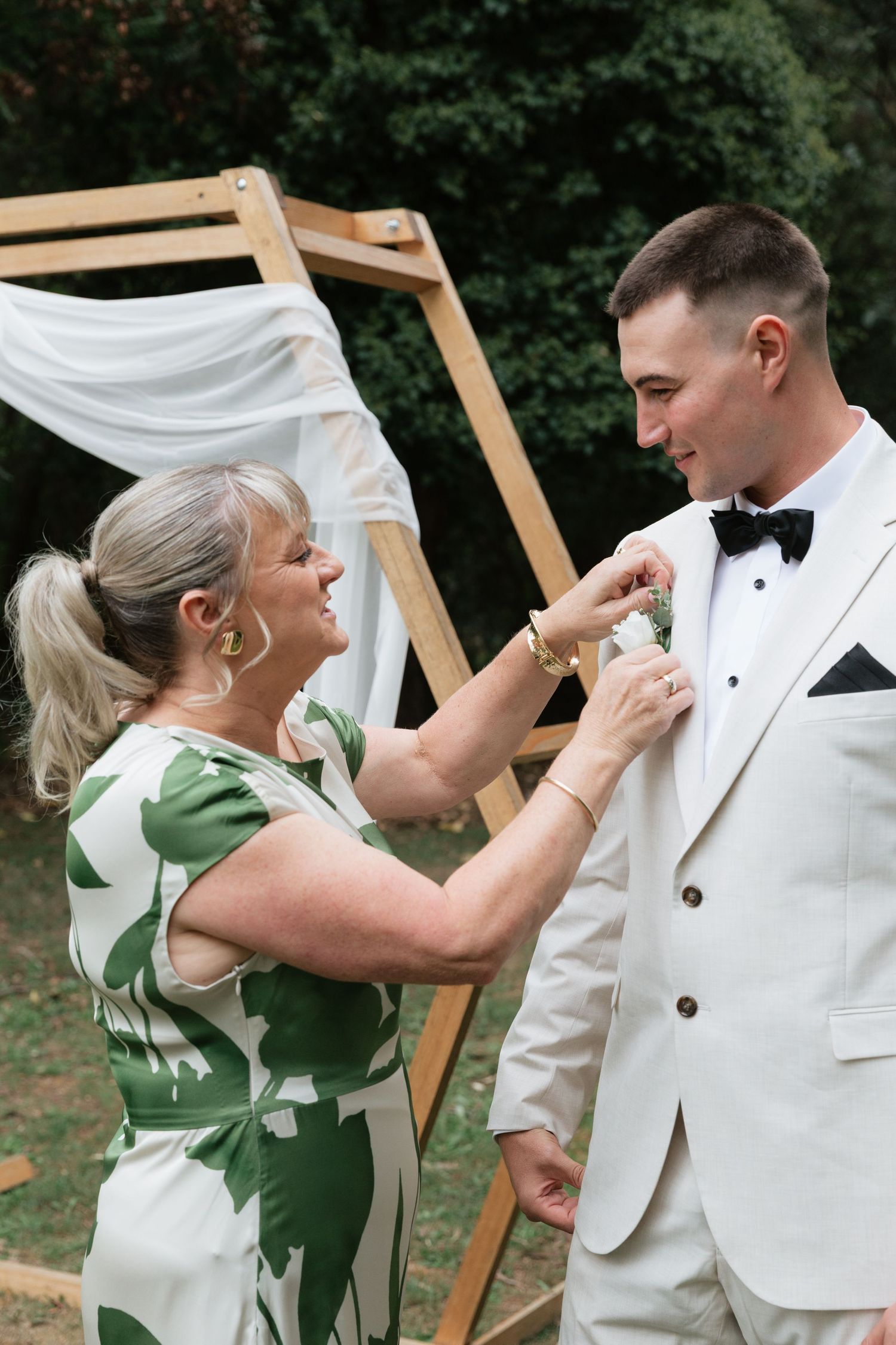 A woman in a green dress pins a boutonniere on a groom in a white tuxedo at an outdoor wedding ceremony.