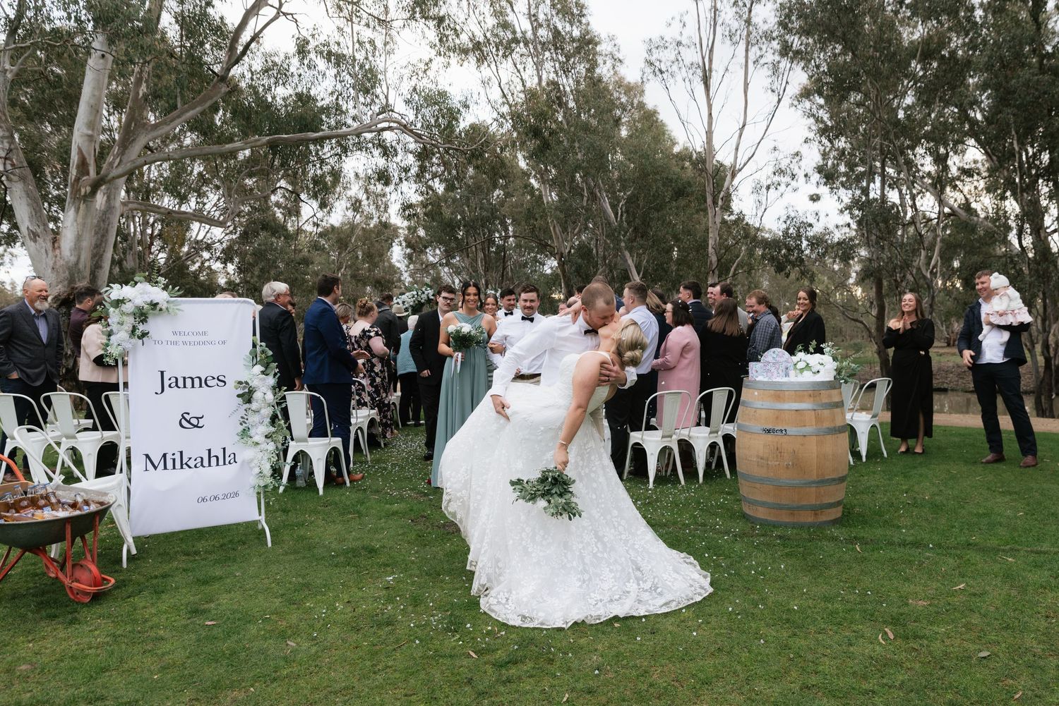 Bride and groom kiss at outdoor wedding ceremony with white chairs and barrel decor under eucalyptus trees.