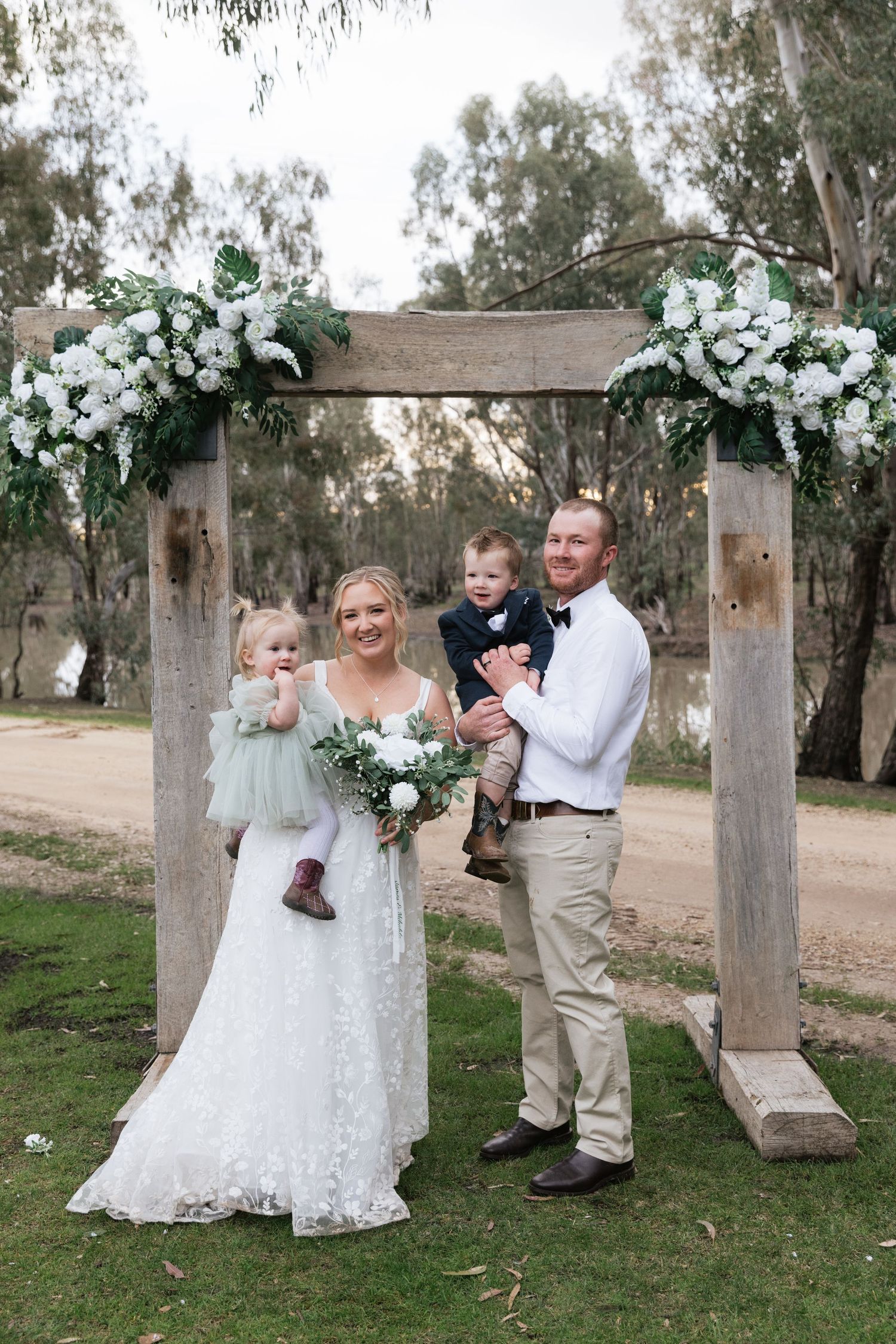 Bride and groom stand under floral arch holding two young children at outdoor wedding ceremony near river.