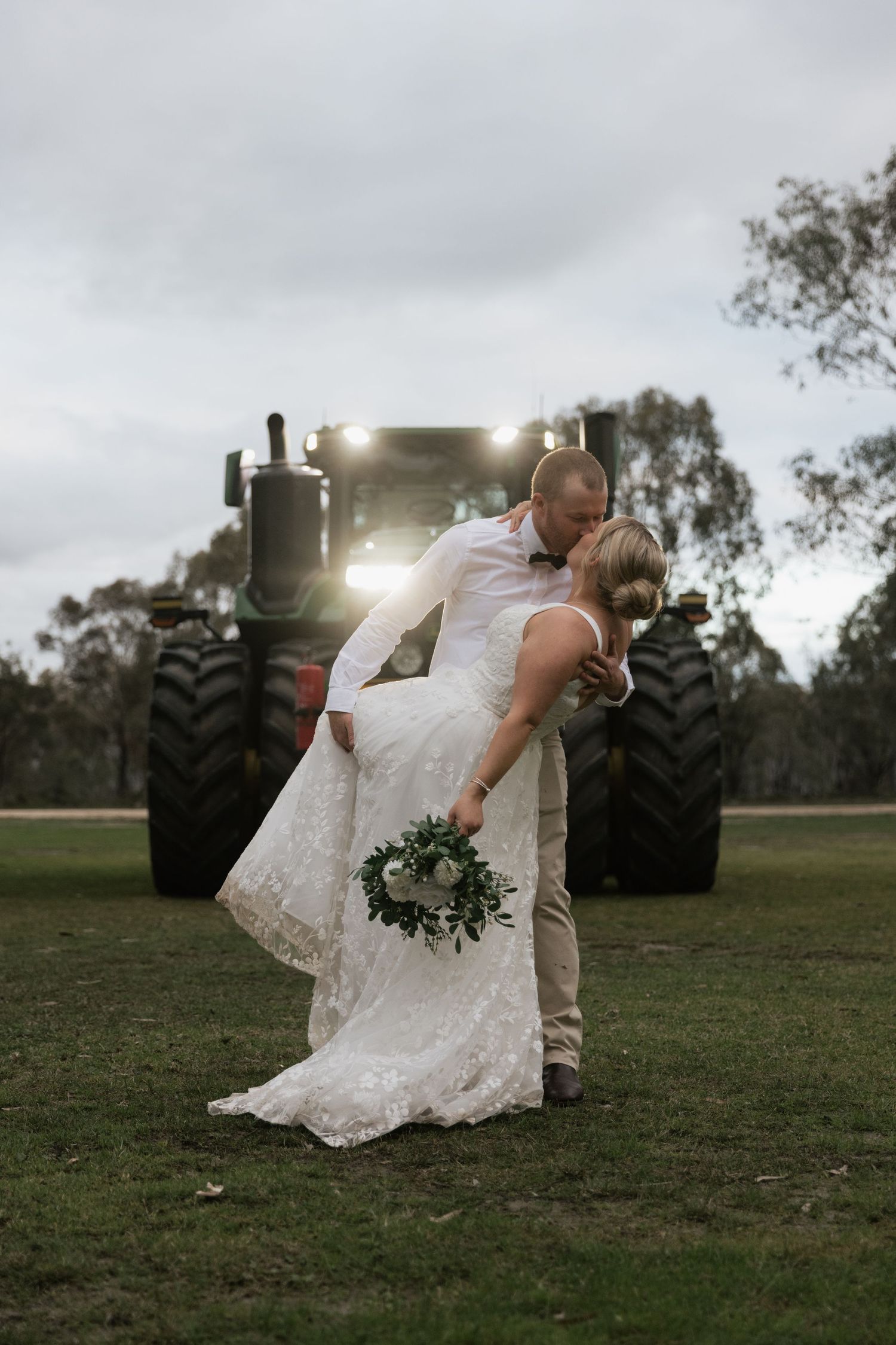 A bride and groom share a romantic dip kiss in front of a large tractor on a farm at dusk, bouquet in hand.