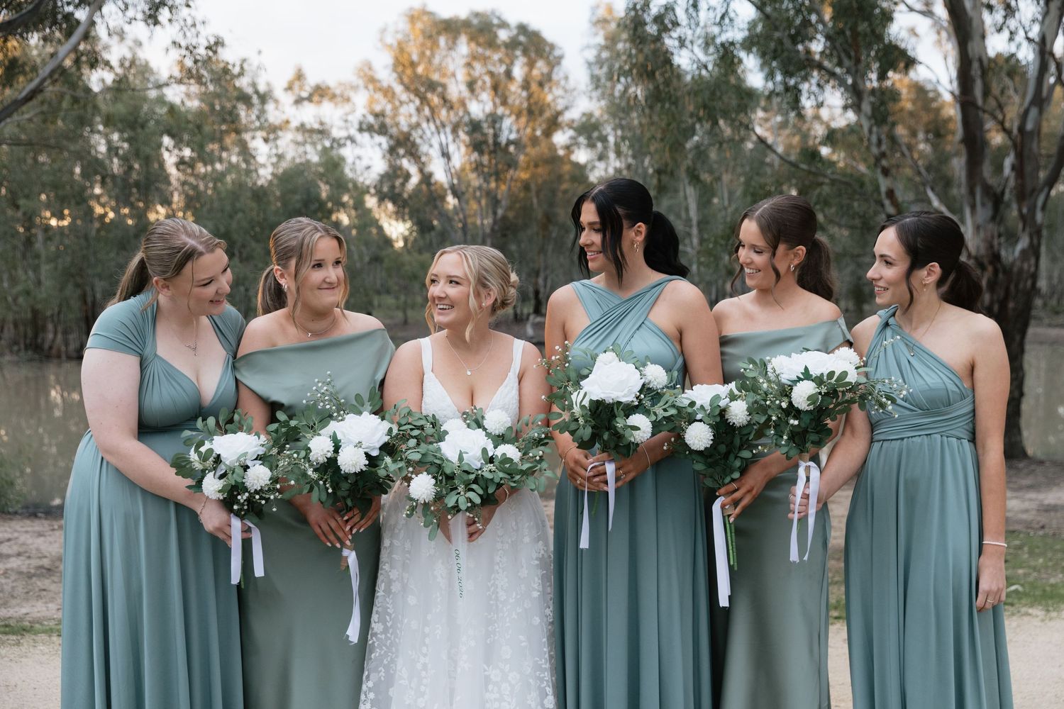 Bride in white lace gown with five bridesmaids in sage green dresses holding white floral bouquets outdoors.