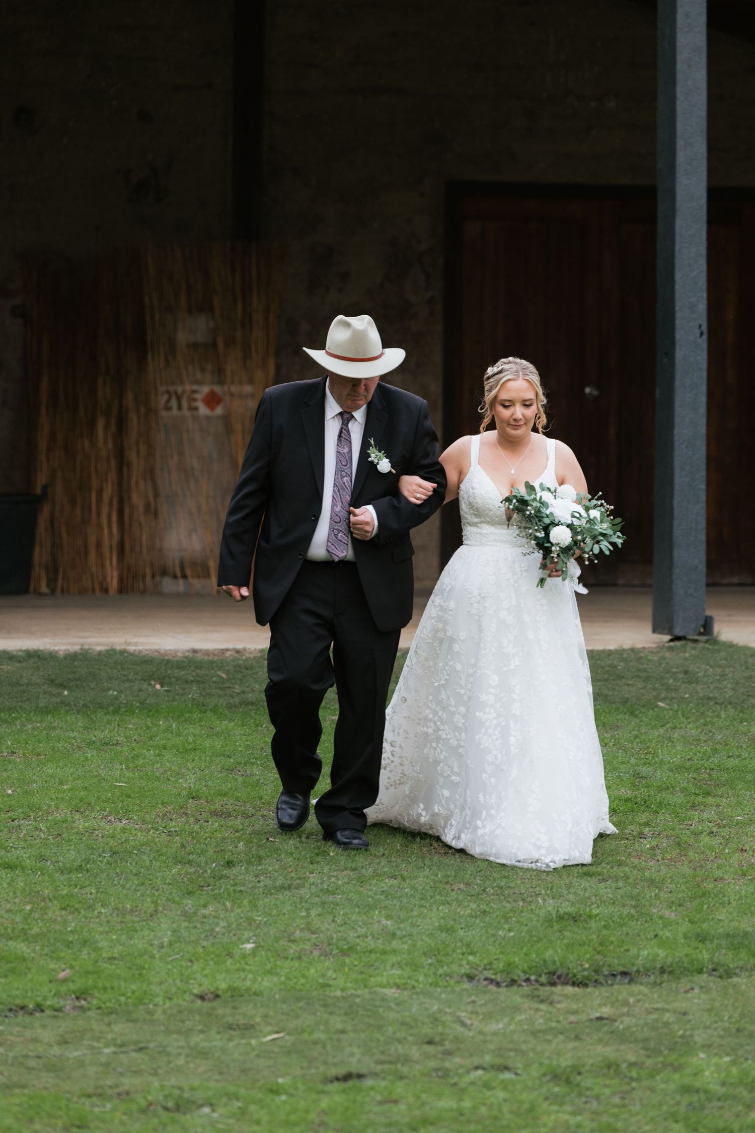 Father walks bride in white lace gown down the aisle at outdoor rustic wedding ceremony.