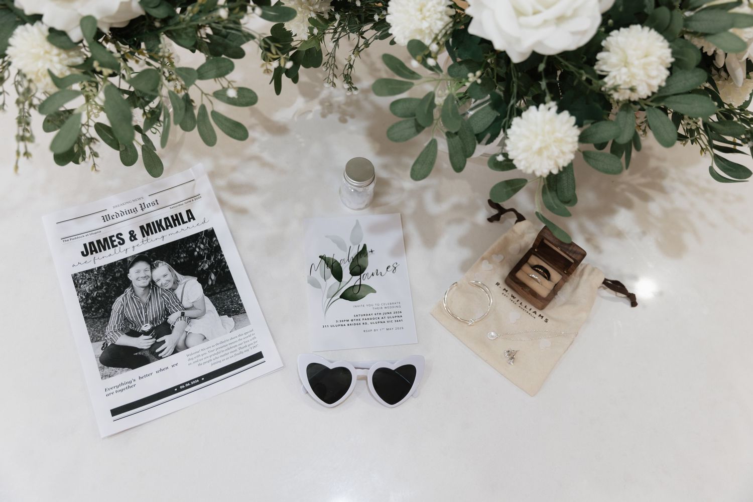Wedding flat lay with floral arrangement, heart sunglasses, rings, and James & Mikayla ceremony programs on white background.