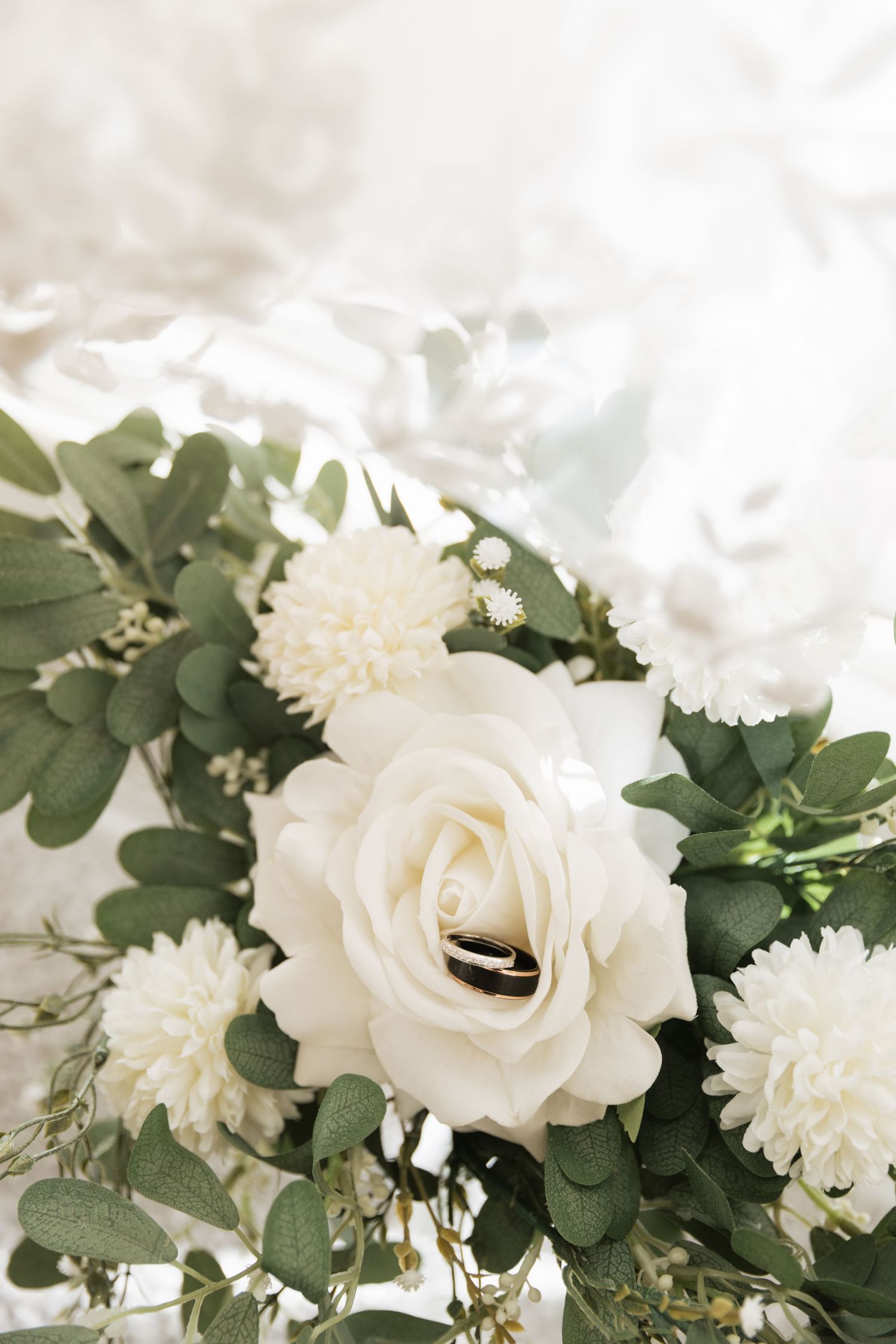 White rose bouquet with gold wedding rings, green eucalyptus leaves, and white chrysanthemums on a soft bridal veil.