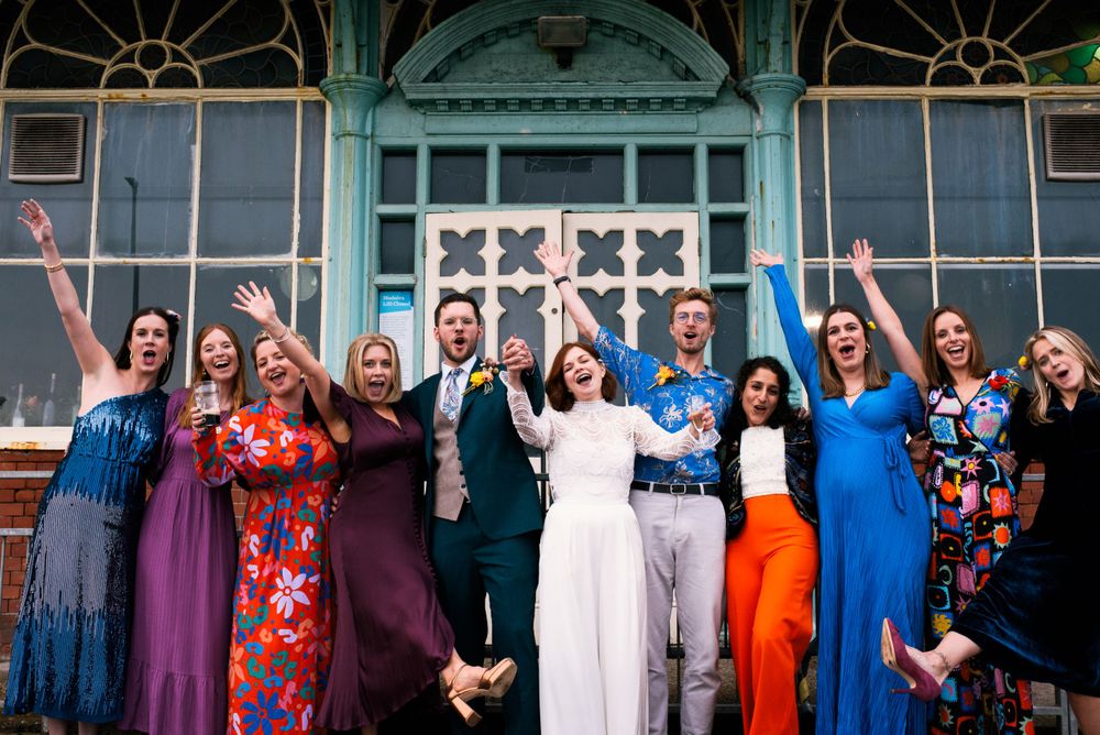 Wedding party poses in front of turquoise Victorian architecture wearing colorful attire.