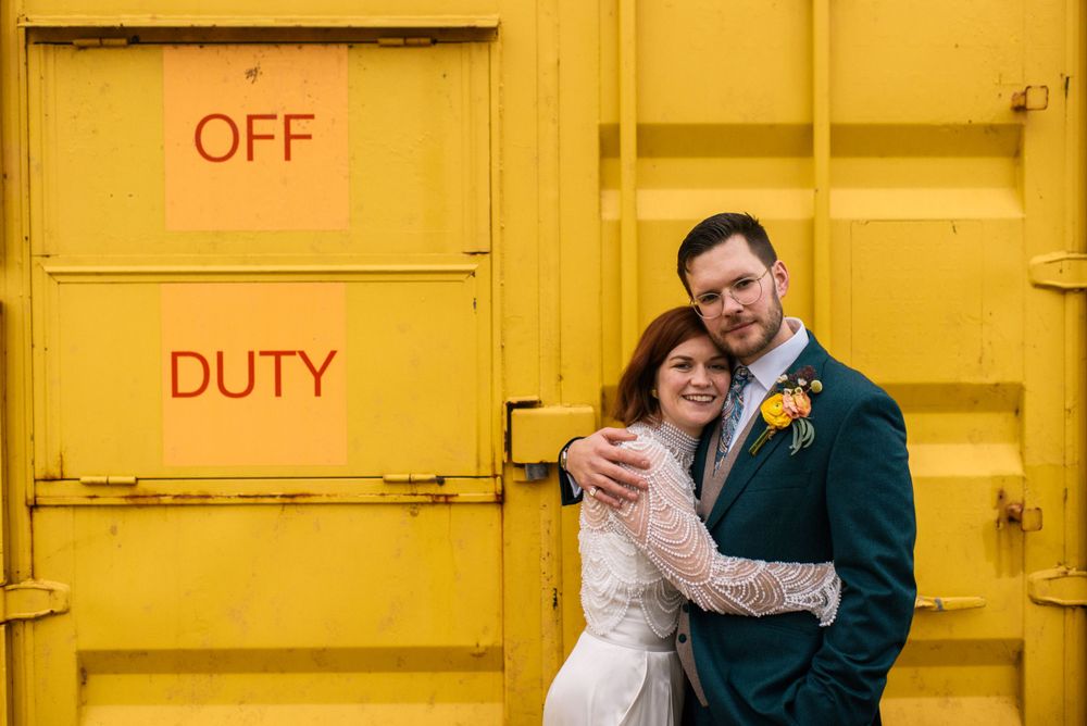 Newlyweds share intimate moment against bright yellow industrial door marked 'Off Duty'.