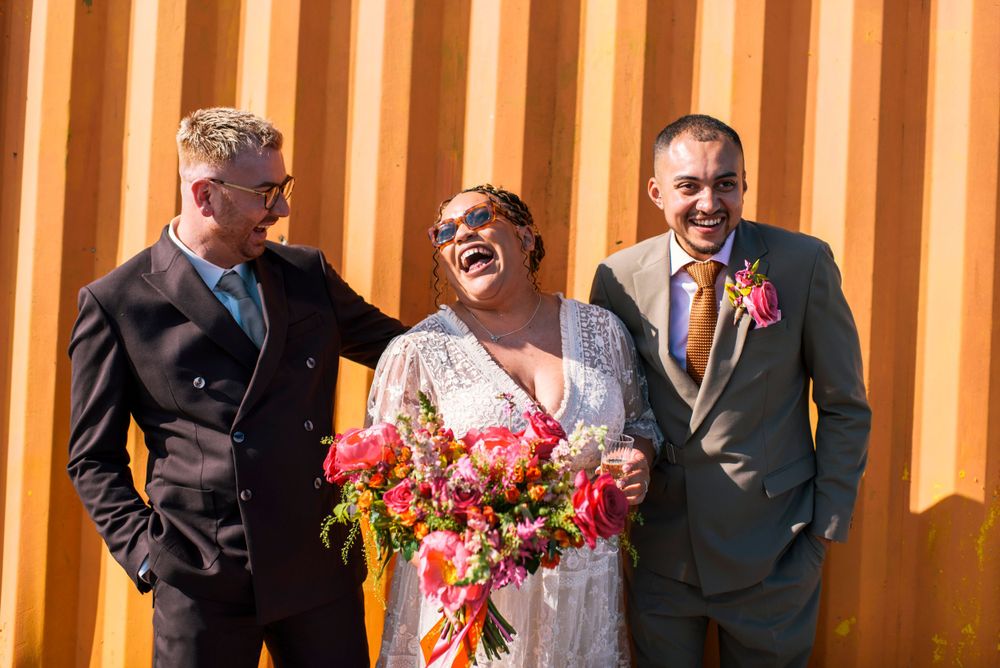 Wedding party members share laughter against vibrant orange wall with bright floral bouquet.