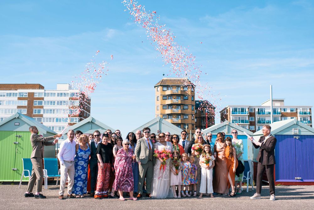 Wedding guests gather for group photo in front of colorful beach huts as confetti rains down.