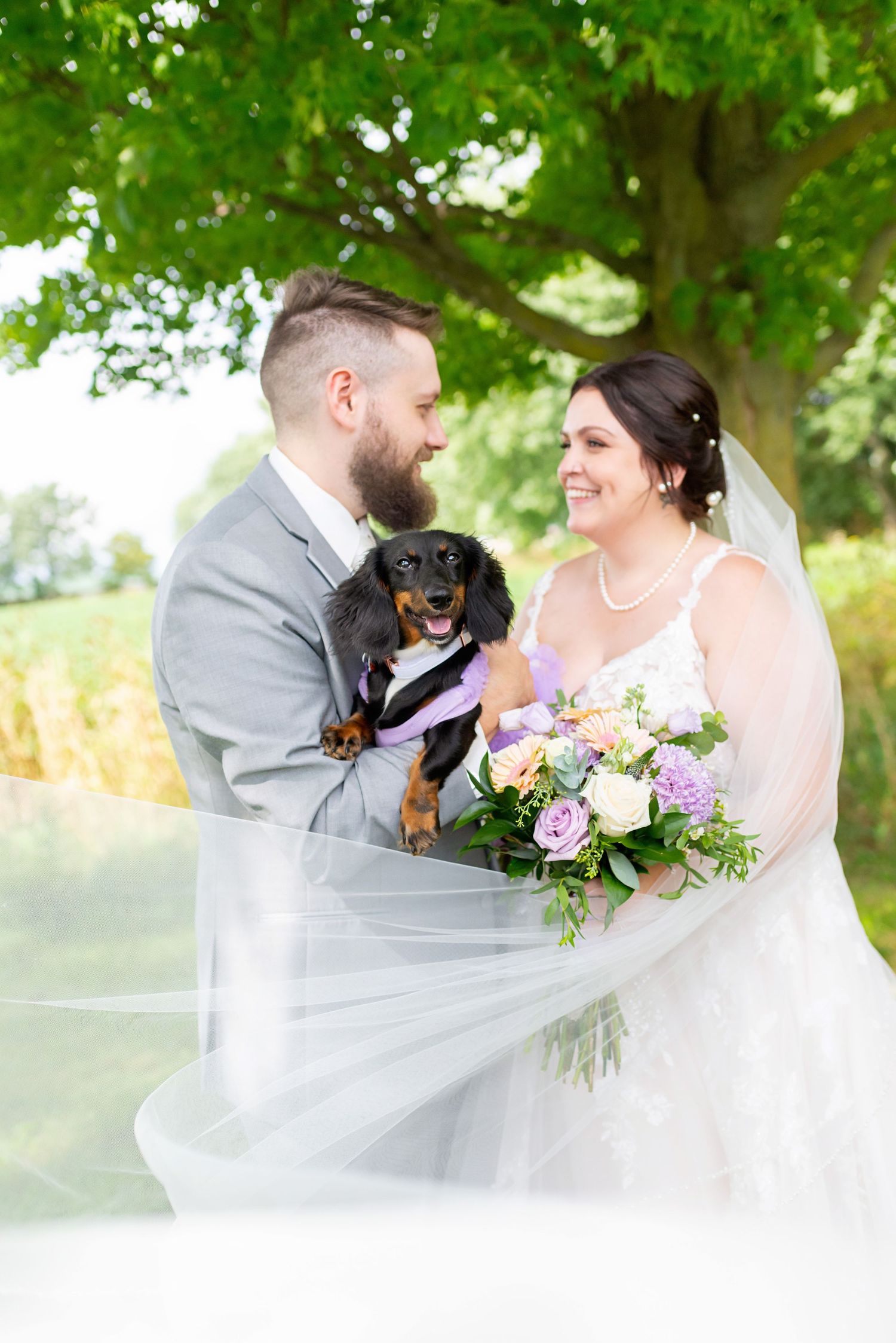 Wedding couple sharing intimate moment under green trees with floral bouquet in Strathroy at Mac Cuddy Botanic Garden.