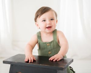 Baby in green overalls leans on dark wooden box in bright white room.