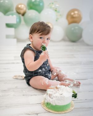 Baby in dark romper enjoying first birthday cake smash with mint green decorations.