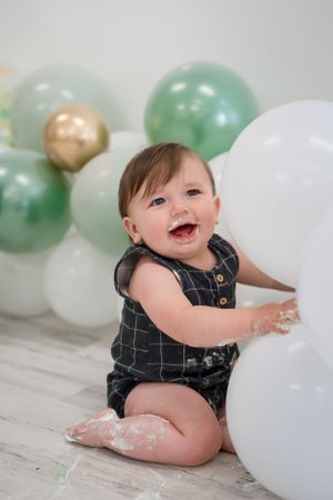 Baby in dark romper plays with balloons on wooden floor with mint and gold balloon backdrop.
