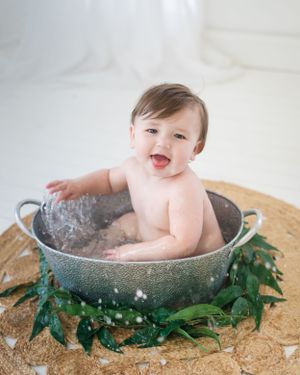 Baby splashes in vintage metal tub decorated with greenery during bath photoshoot.