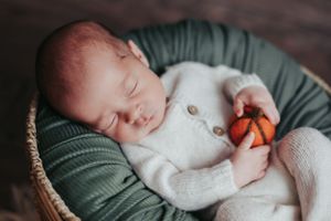 Sleeping newborn in white sweater holding tiny pumpkin in basket.