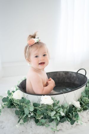 Baby sits in metal tub surrounded by eucalyptus leaves and white roses during photo session.