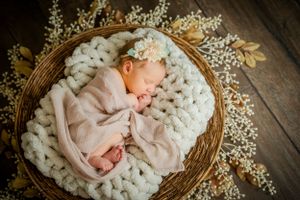 Newborn baby sleeping peacefully in a round basket with white chunky knit blanket and floral accents.