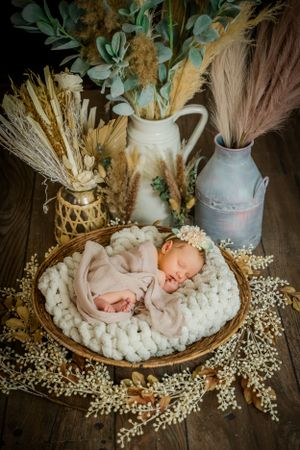 Rustic newborn photography setup with baby in basket surrounded by dried flowers and vintage milk jugs.