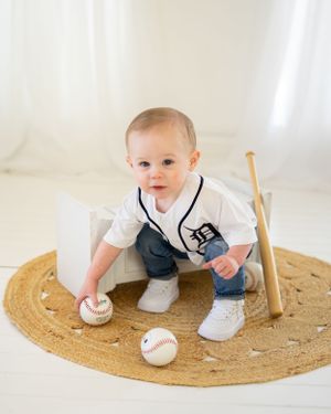 Baby in baseball uniform sits on round jute rug with baseballs and bat.