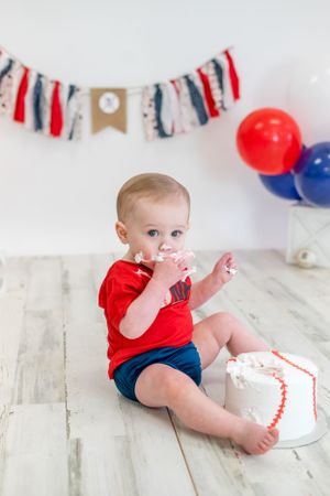 Baby in red shirt and blue shorts with patriotic decorations and cake during celebration.