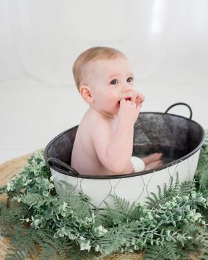 Baby sits in vintage metal tub surrounded by fresh greenery and ferns.