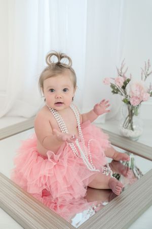 A baby wearing a pink tulle tutu dress and pearl necklace sits on a mirrored surface near pink flowers in a vase.