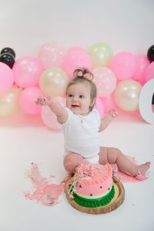 A cake smash photo series showing a baby with a watermelon-themed cake against a pink and white balloon backdrop.