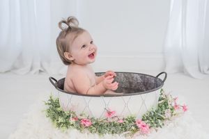 A baby sits in a vintage bathtub surrounded by delicate pink flowers and greenery in a bright white room.
