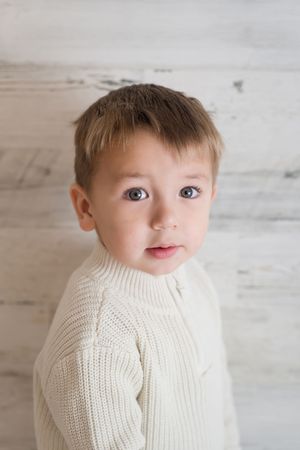 Young child wearing a cozy cream-colored knit sweater poses against a light rustic wooden background.