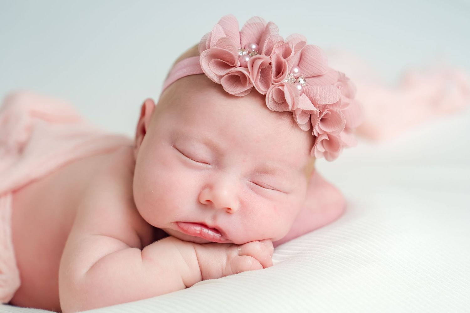 Peaceful sleeping newborn baby wearing pink floral headband resting on soft white blanket.