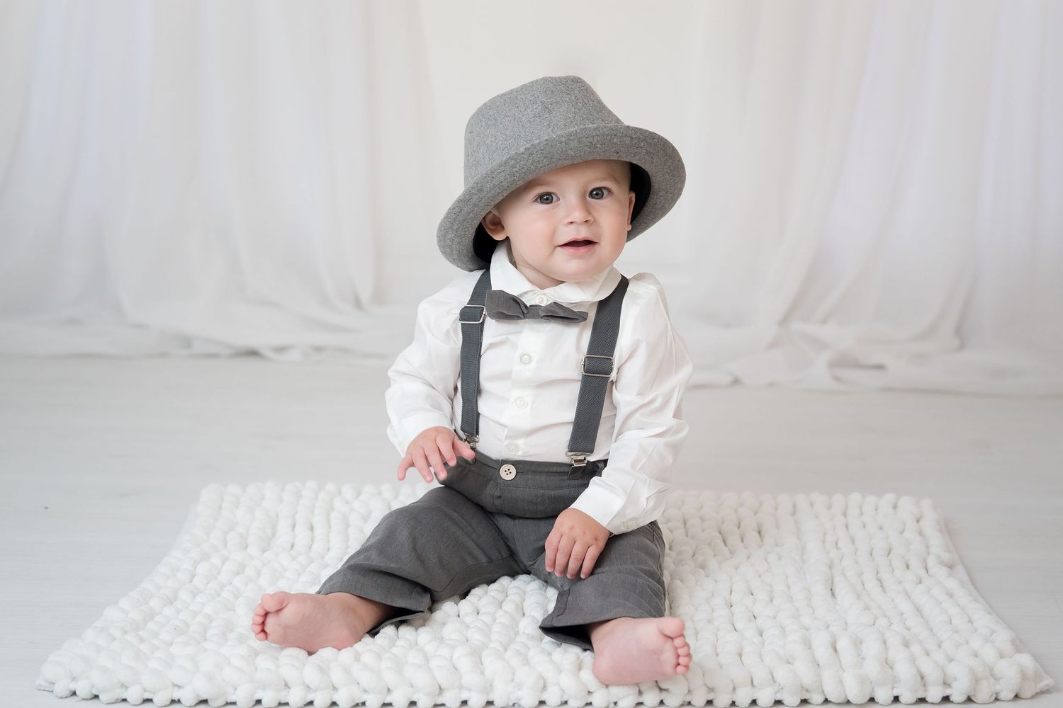 A baby in a gray fedora hat and suspenders sits on a white knit blanket during a photo session.