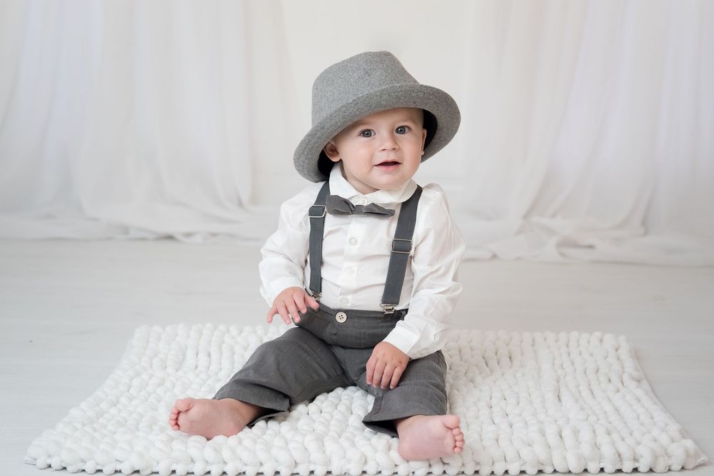 A baby in a gray fedora hat and suspenders sits on a white knit blanket during a photo session.