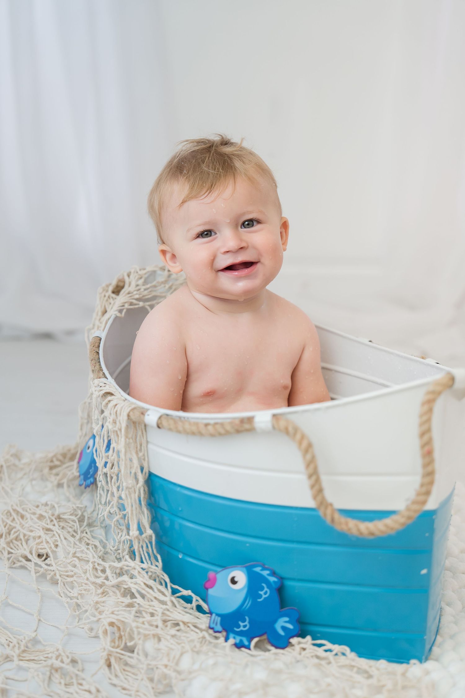 Baby sits in a blue and white decorative boat prop with fishing net and toy fish for nautical themed photo shoot.