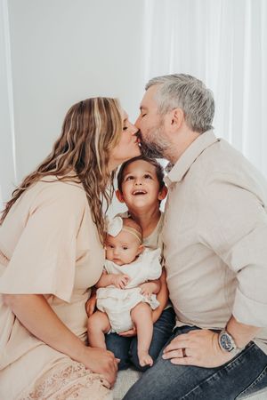 A loving family moment as parents share a kiss while holding their two young children in a bright indoor setting.