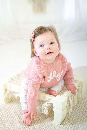 Baby in pink sweater sitting on vintage furniture against white backdrop.