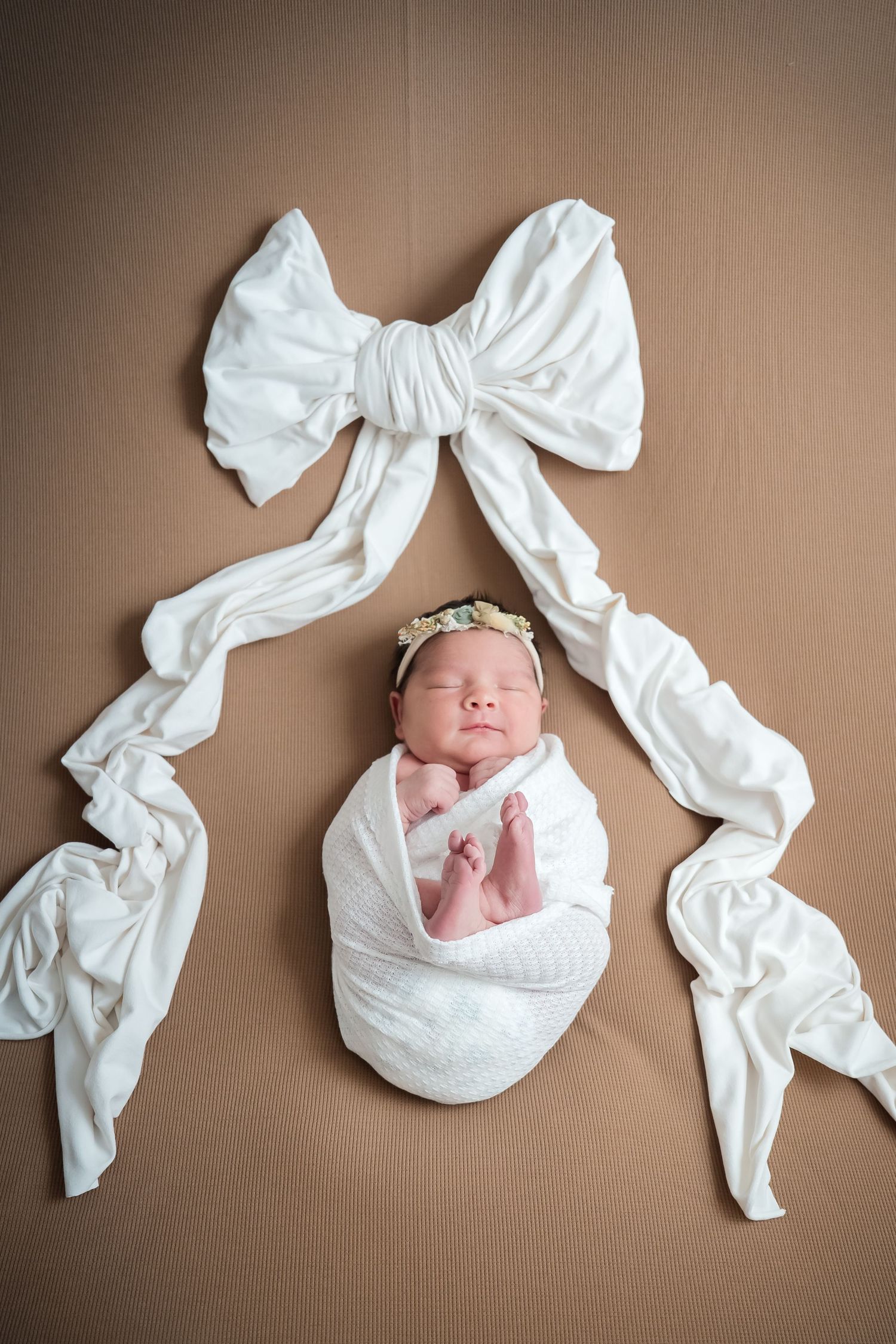 A sleeping newborn baby wrapped in white fabric with decorative bow arrangements on a brown backdrop.